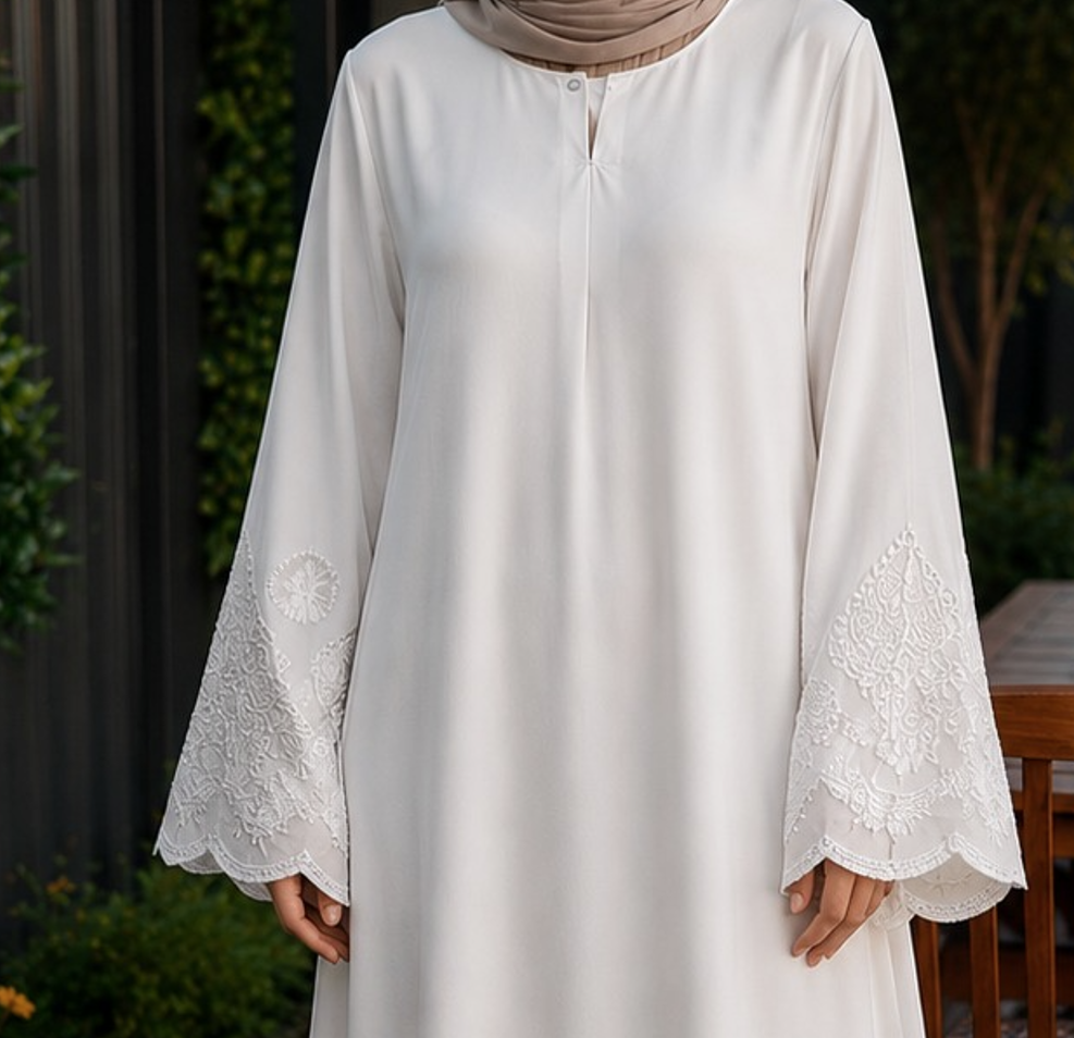 A woman wearing a long white abaya dress with wide bell sleeves and intricate floral lace embroidery at the cuffs, paired with a beige hijab, photographed outdoors against a dark fence and greenery.