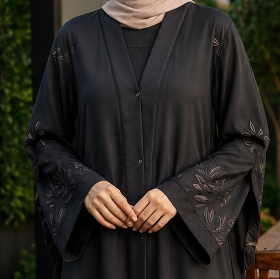 A woman wearing a dark charcoal black button-front abaya with tonal floral embroidery on the sleeves, paired with a light beige hijab. The background is a soft-focus outdoor setting with green foliage.