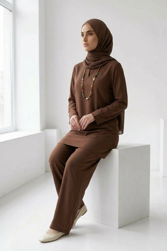 A woman wearing a matching brown long-sleeve tunic and wide-leg pants set with a coordinating hijab, sitting in a bright white studio with geometric display blocks.