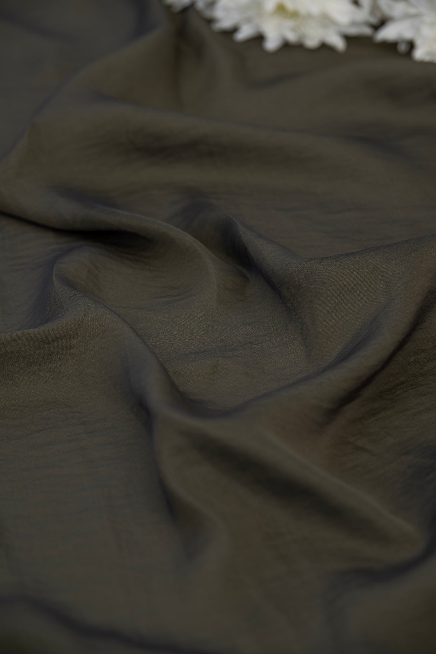 A close-up of draped dark olive green fabric with soft folds and a small cluster of white flowers at the top edge.
