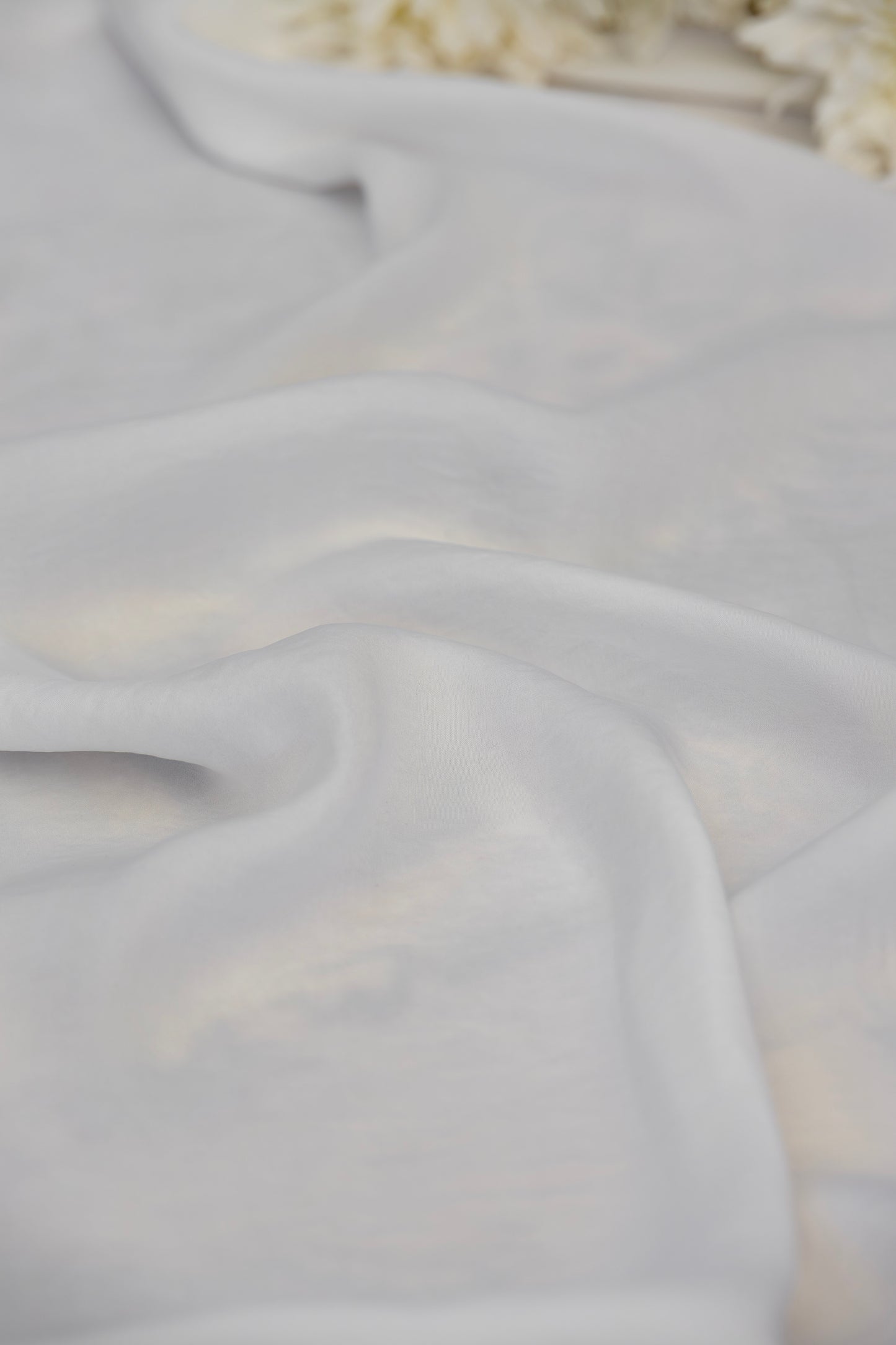 A close-up of draped white fabric with soft folds and a subtle sheen, with a blurred decorative floral element in the top right corner.