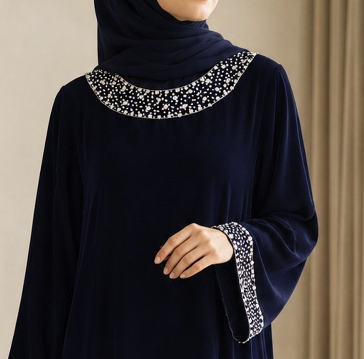 A woman wearing a long white abaya dress with wide bell sleeves and intricate floral lace embroidery at the cuffs, paired with a beige hijab, photographed outdoors against a dark fence and greenery.
