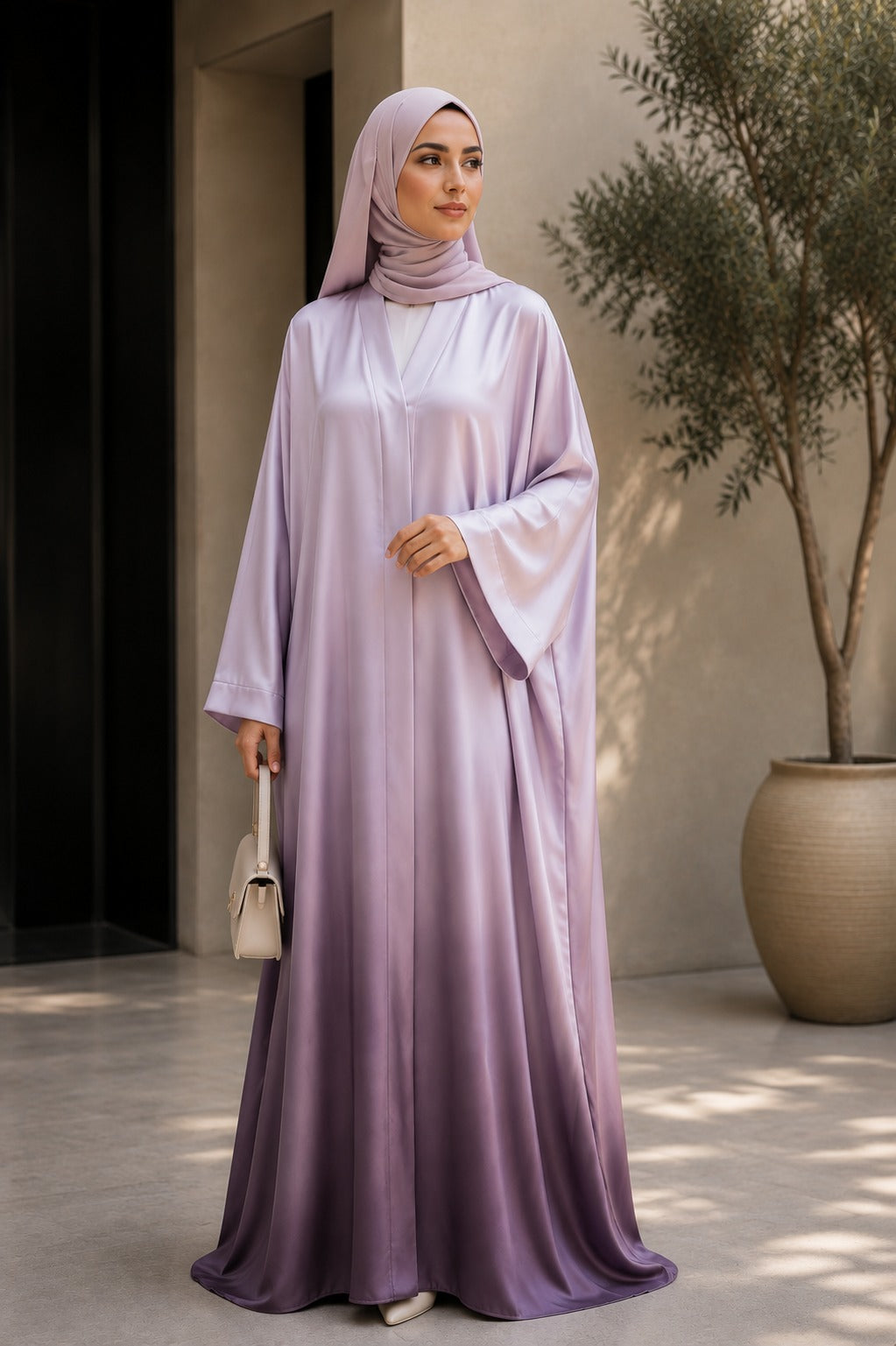 A woman wearing a flowing lavender abaya with a matching hijab, standing in a sunlit courtyard with a large potted tree and neutral-toned walls in the background.