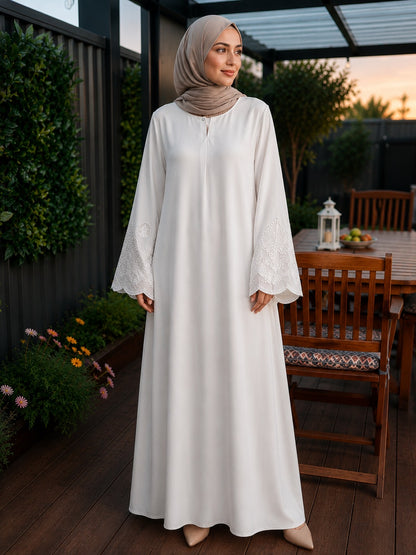 A woman wearing a long white modest maxi dress with lace-trimmed bell sleeves and a taupe hijab, standing on a wooden patio at dusk.