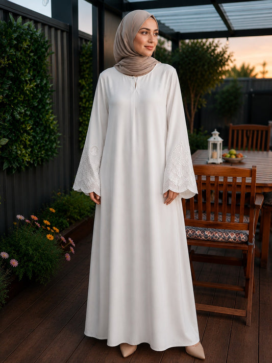 A woman wearing a long white modest maxi dress with lace-trimmed bell sleeves and a taupe hijab, standing on a wooden patio at dusk.