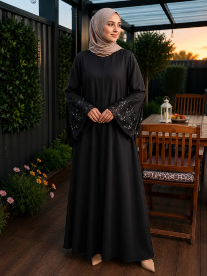 A woman wearing a black long abaya with embroidered bell sleeves and a taupe hijab, standing on a wooden deck in a backyard patio setting.