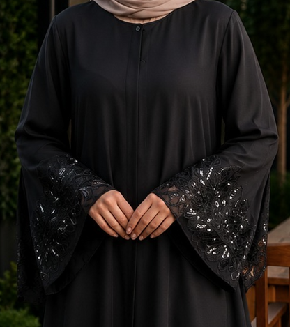 A person wearing a black long-sleeve abaya with wide bell sleeves featuring black lace and sequin embellishments, paired with a beige hijab. The background shows a softly blurred outdoor garden setting.