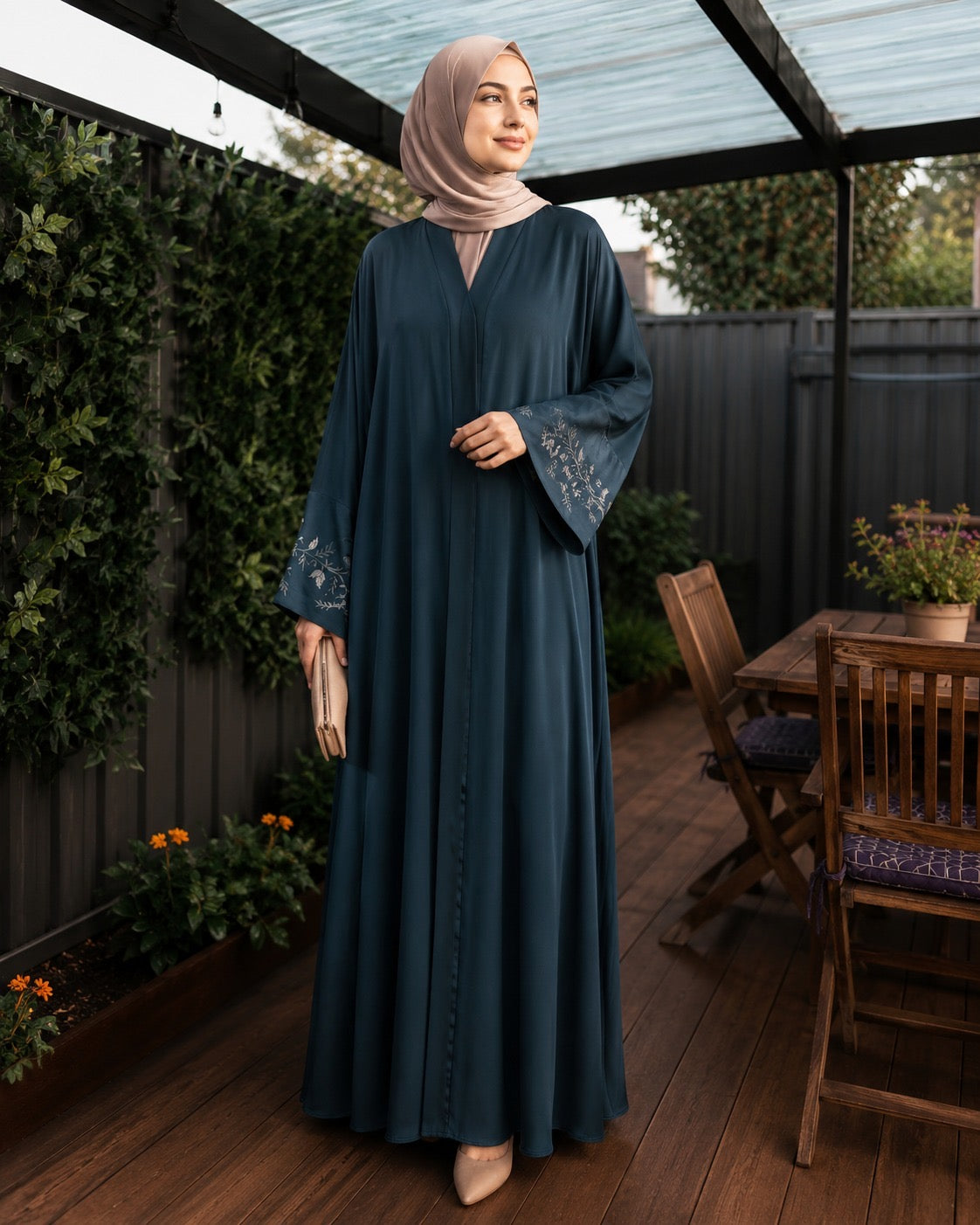 A woman wearing a long teal abaya with embroidered cuffs and a light pink hijab, standing on a wooden deck patio with a garden background.