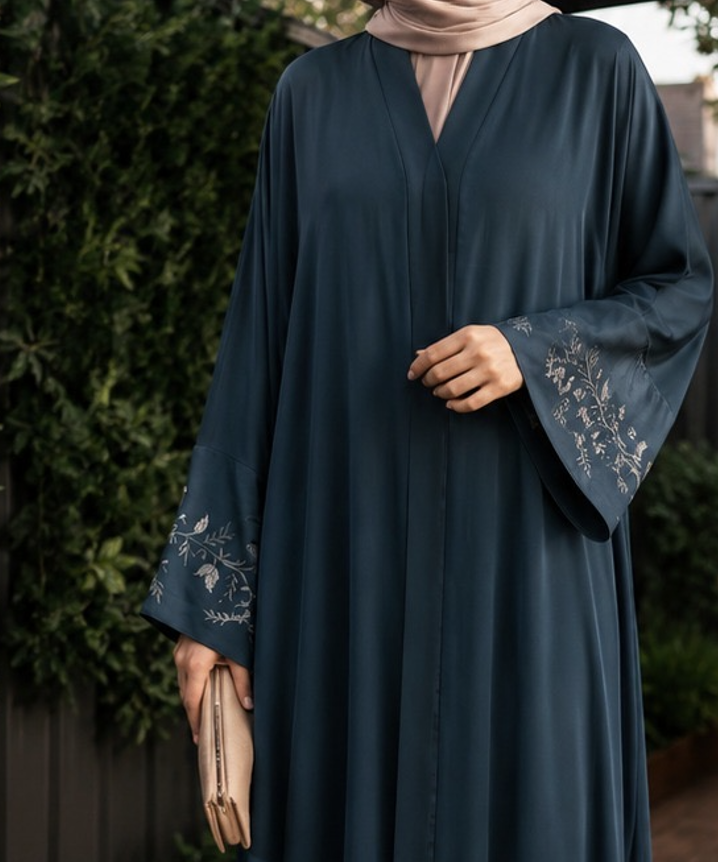 A woman wearing a dark teal abaya with silver floral embroidery on the wide sleeves, paired with a light beige hijab and holding a beige clutch, set against a green hedge background.