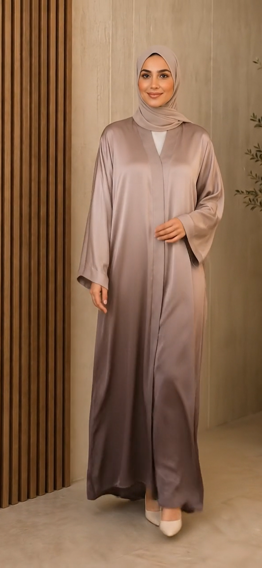 A woman wearing a long, flowing abaya in a dusty mauve-purple color with a matching hijab, standing in front of a modern wooden slatted wall.