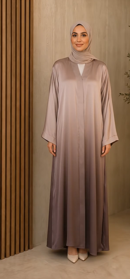 A woman wearing a long, floor-length abaya dress in a dusty mauve-brown color with a subtle sheen, paired with a matching hijab and beige pointed shoes, standing against a modern wooden-paneled background.