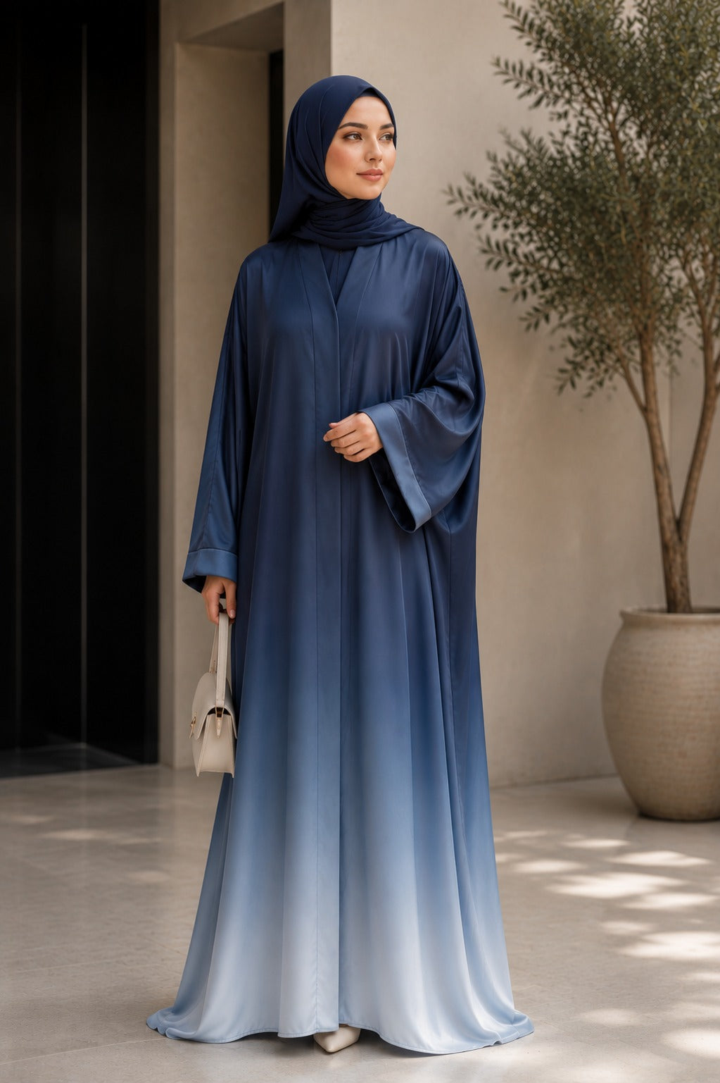 A woman wearing a long blue-to-light-blue gradient abaya with a matching hijab, standing in a modern indoor setting with a potted tree and neutral walls.