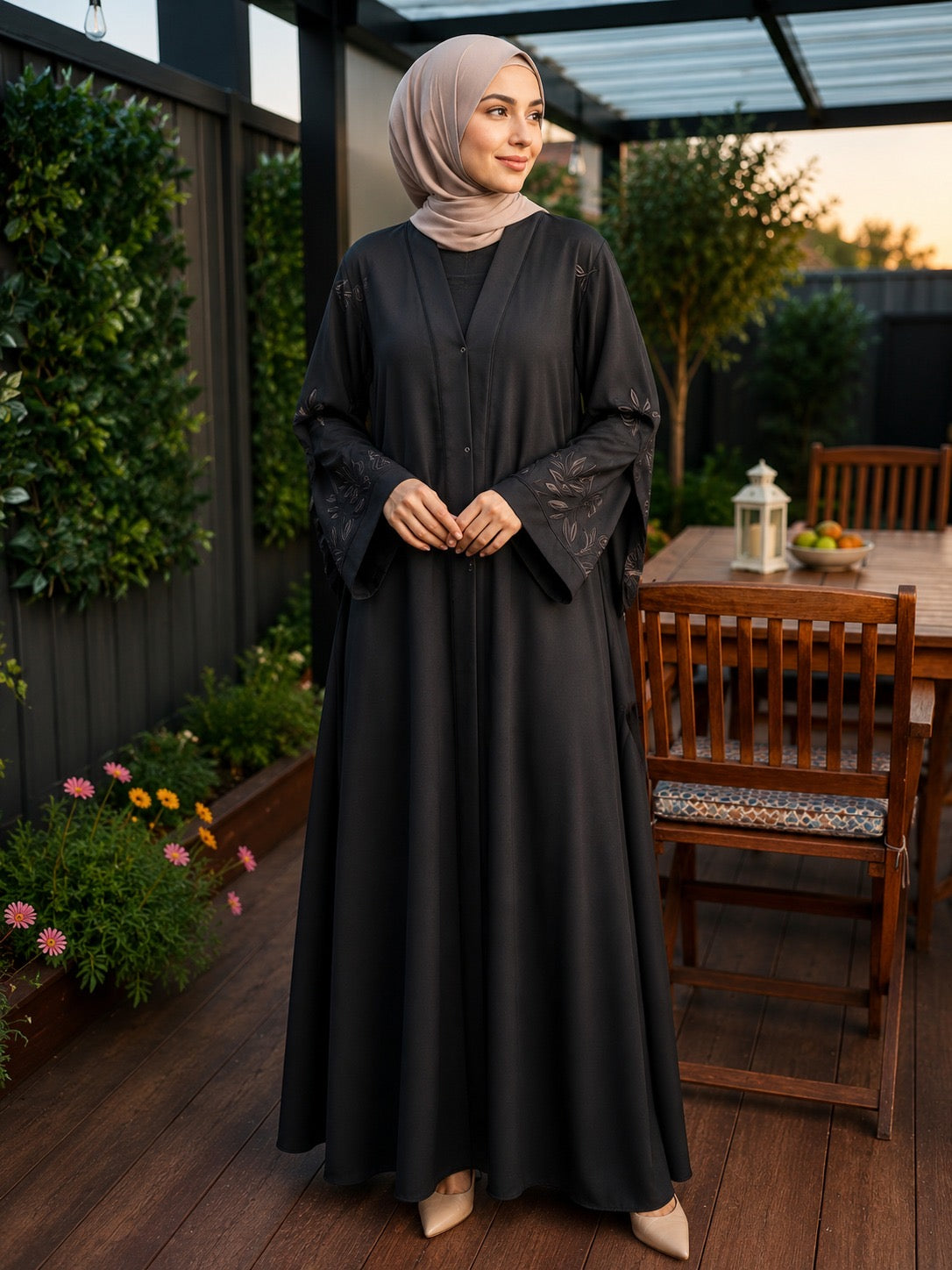 A woman wearing a black long abaya with sheer embroidered sleeves and a matching light taupe hijab, standing on a wooden patio with garden plants and a dining table in the background.