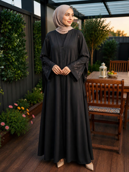 A woman wearing a black long abaya with sheer embroidered sleeves and a matching light taupe hijab, standing on a wooden patio with garden plants and a dining table in the background.