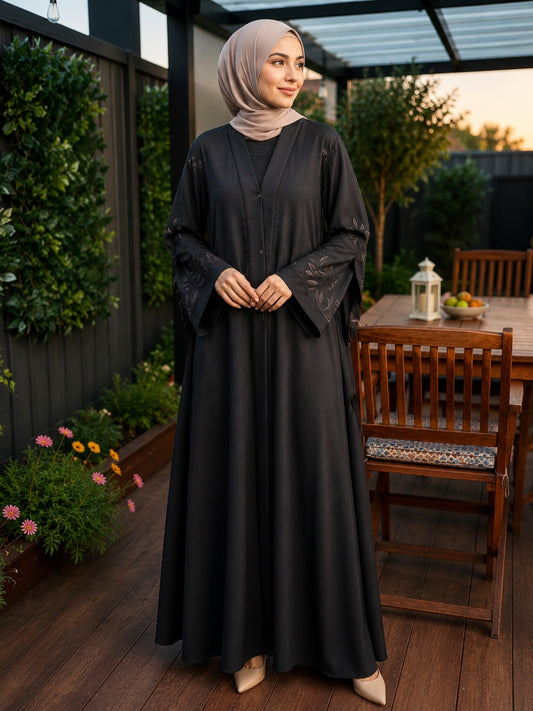 A woman wearing a black long abaya with sheer embroidered sleeves and a matching light taupe hijab, standing on a wooden patio with garden plants and a dining table in the background.