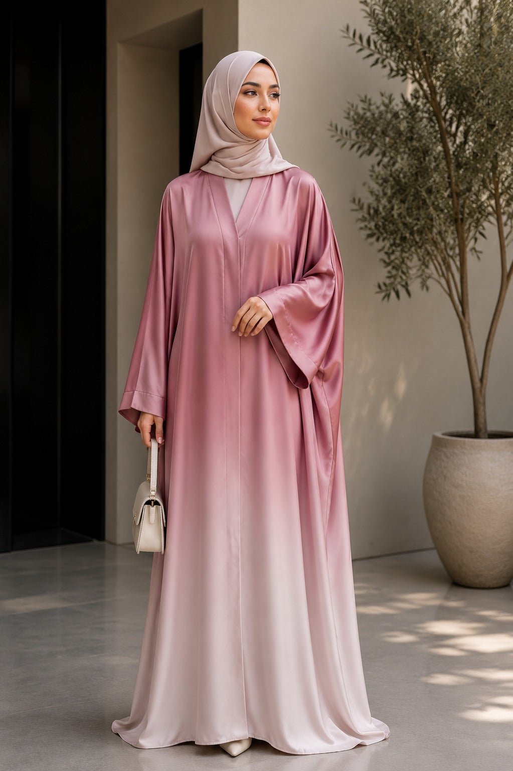A woman wearing a long, flowing dusty pink abaya with a gradient effect and a matching light pink hijab, standing in a modern indoor setting with a potted tree.