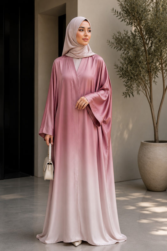 A woman wearing a long, flowing dusty pink abaya with a gradient effect and a matching light pink hijab, standing in a modern indoor setting with a potted tree.