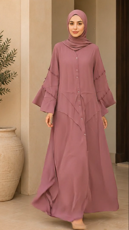 A woman wearing a dusty rose abaya dress and matching hijab, standing in an indoor setting with beige walls and a large ceramic pot.