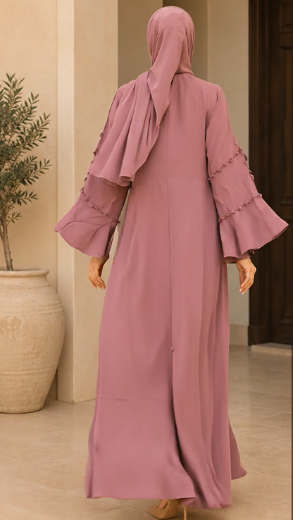 A woman wearing a dusty pink abaya with a matching hijab, shown from the back in an indoor setting with a beige wall and potted plant.