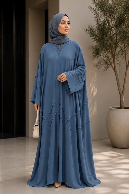 A woman wearing a flowing slate blue abaya and matching hijab, standing in a modern indoor setting with a potted tree and neutral walls.