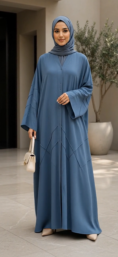 A woman wearing a long, flowing blue abaya with a matching high-neck hijab, standing in a modern indoor setting with a potted plant in the background.