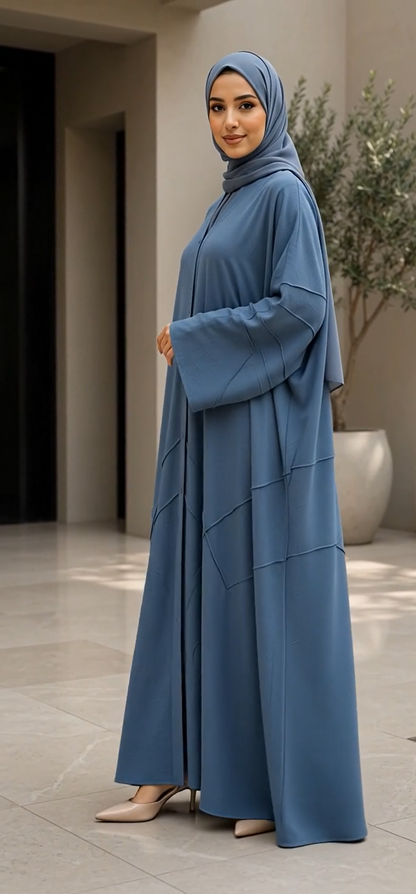 A woman wearing a modest blue abaya and matching hijab stands in a modern indoor setting with neutral walls and a potted plant in the background.