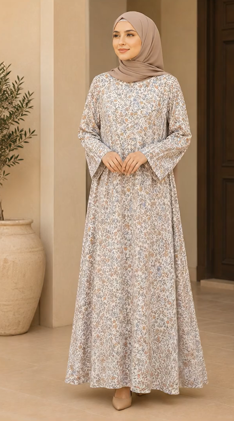 A woman wearing a long-sleeved, floor-length abaya dress with a delicate beige and white floral pattern, paired with a matching taupe hijab, standing in a neutral-toned indoor setting.