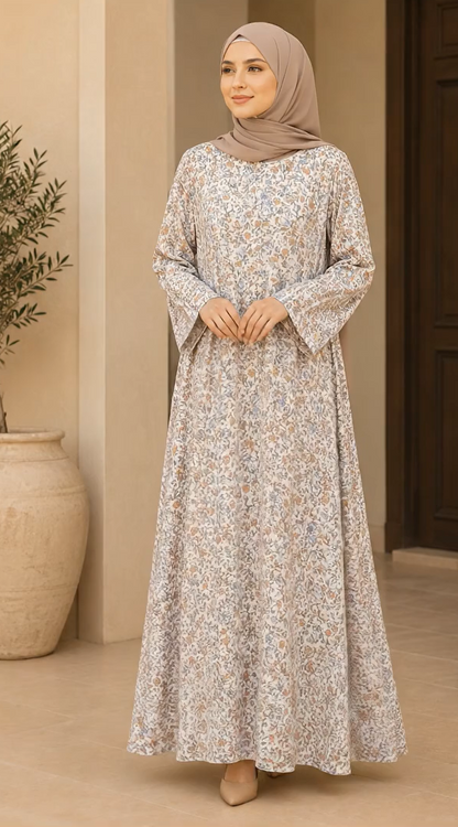 A woman wearing a long-sleeved, floor-length abaya dress with a delicate beige and white floral pattern, paired with a matching taupe hijab, standing in a neutral-toned indoor setting.