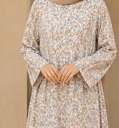 A woman wearing a long-sleeved, V-neck modest dress with a dense floral and botanical print in beige, light blue, and soft brown tones, set against a solid beige background.