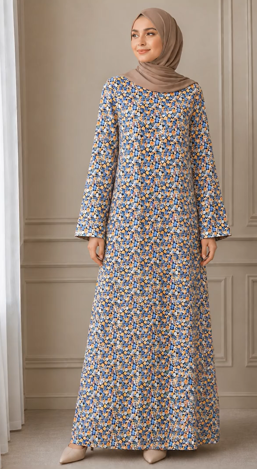 A woman wearing a long-sleeved maxi abaya dress with a dense blue, yellow, and white floral print, paired with a taupe hijab, standing in front of a beige paneled wall.