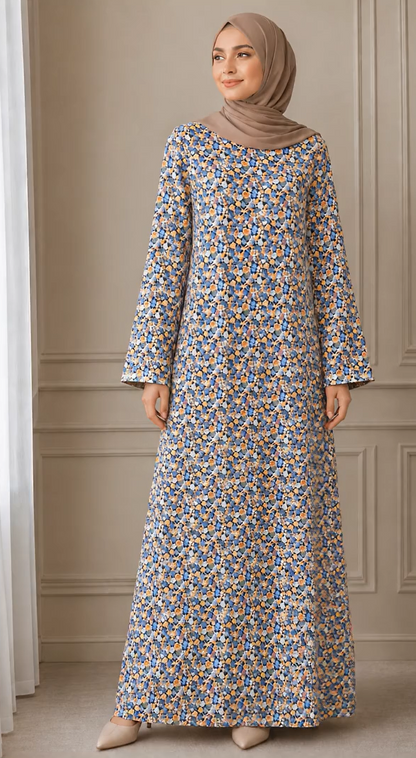 A woman wearing a long-sleeved maxi abaya dress with a dense blue, yellow, and white floral print, paired with a taupe hijab, standing in front of a beige paneled wall.