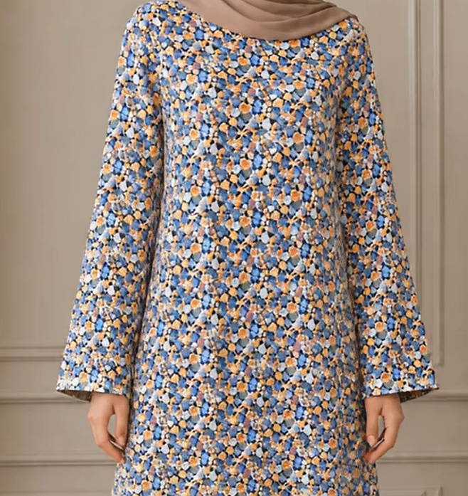 A woman wearing a long-sleeved tunic dress with a dense blue, orange, and white floral pattern, paired with a solid taupe hijab. The background is a soft beige paneled wall.