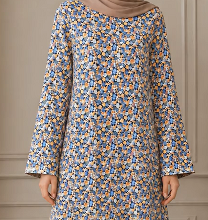 A woman wearing a long-sleeved tunic dress with a dense blue, orange, and white floral pattern, paired with a solid taupe hijab. The background is a soft beige paneled wall.