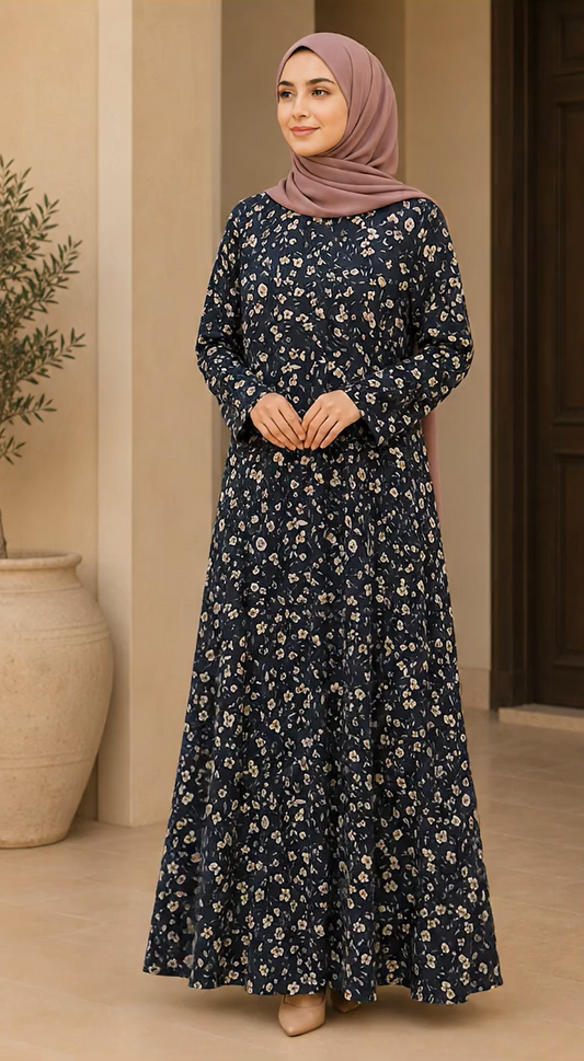 A woman wearing a long-sleeved navy blue maxi dress with a small white floral print and a dusty rose hijab, standing in an indoor setting with beige walls and a large ceramic pot.