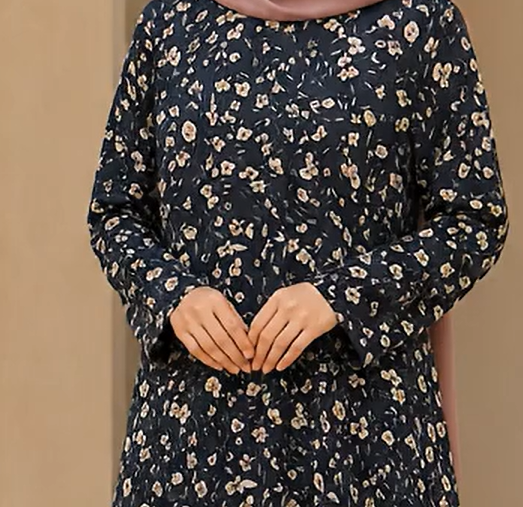 A woman wearing a navy blue long-sleeve dress with a dense beige and white floral print, paired with a dusty pink hijab. The photo is cropped from the chest down, set against a plain beige background.