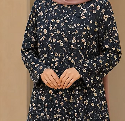A woman wearing a navy blue long-sleeve dress with a dense beige and white floral print, paired with a dusty pink hijab. The photo is cropped from the chest down, set against a plain beige background.