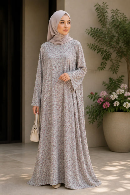 A woman wearing a long-sleeved, floor-length abaya dress with a small floral print in light gray and beige, paired with a matching hijab, standing in a sunlit patio setting.