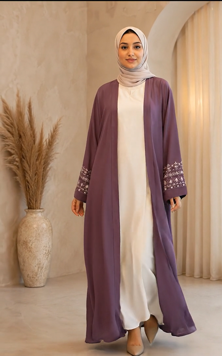 A woman wearing a long, open-front mauve abaya with embroidered cuffs over a white dress and a light beige hijab, standing in a warm-toned indoor setting with a pampas grass vase.