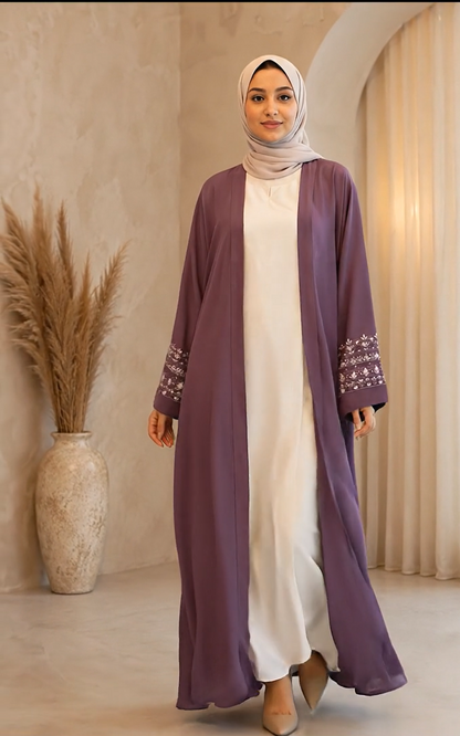 A woman wearing a long, open-front mauve abaya with embroidered cuffs over a white dress and a light beige hijab, standing in a warm-toned indoor setting with a pampas grass vase.