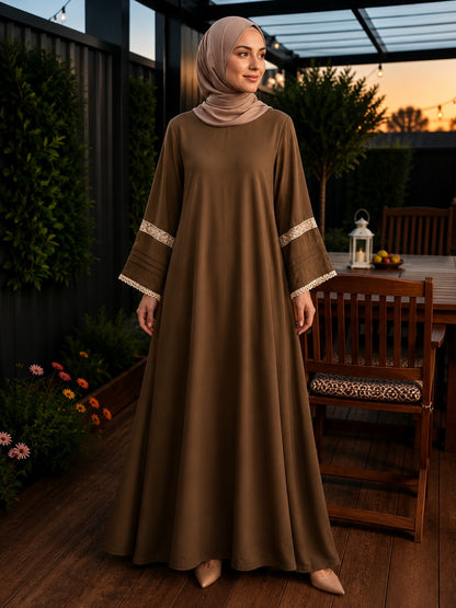 A woman wearing a long brown modest maxi dress with beige lace trim on the sleeves and a matching beige hijab, standing on a wooden deck at sunset.