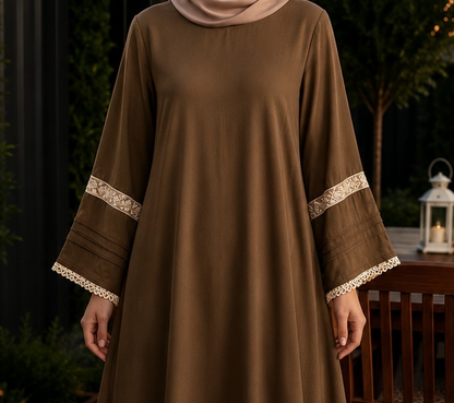 A woman wearing a brown long-sleeve abaya dress with beige lace trim at the cuffs and a matching beige hijab, photographed outdoors at dusk near a wooden railing and lantern.