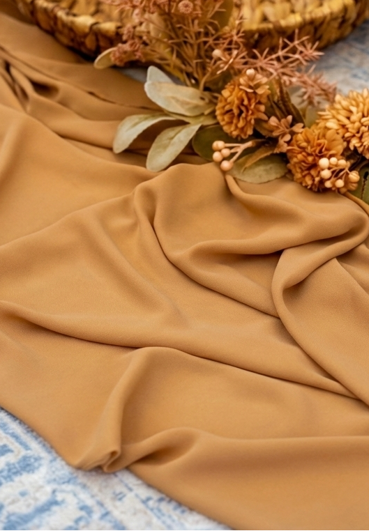 A draped piece of warm camel-colored fabric, styled with dried flowers and a woven basket on a blue patterned textile background.