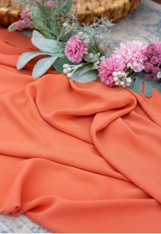 A draped piece of peach-colored fabric with soft folds, styled with pink flowers and green foliage on a patterned blue rug.