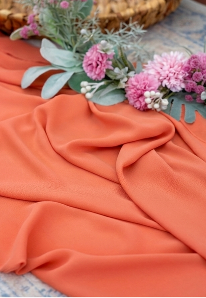 A draped piece of peach-colored fabric with soft folds, styled with pink flowers and green foliage on a patterned blue rug.