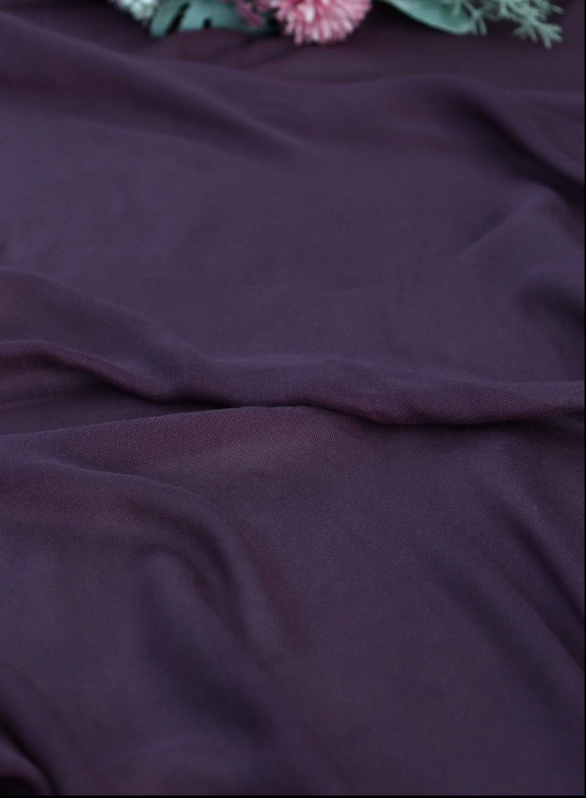 A close-up of draped dark plum purple fabric with soft folds and a small floral prop visible at the top edge.