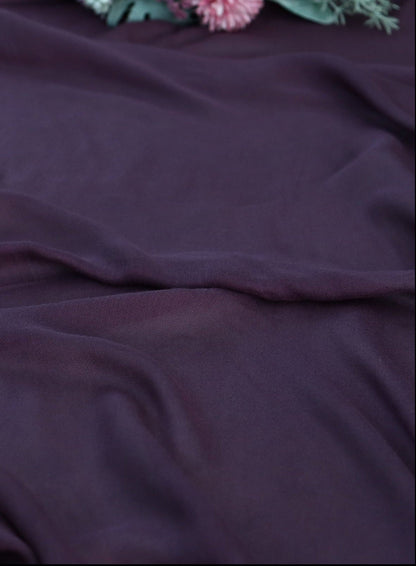 A close-up of draped dark plum purple fabric with soft folds and a small floral prop visible at the top edge.