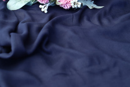 A close-up of draped navy blue fabric with soft folds and a small floral arrangement in the top corner featuring pink blossoms, white berries, and green leaves.