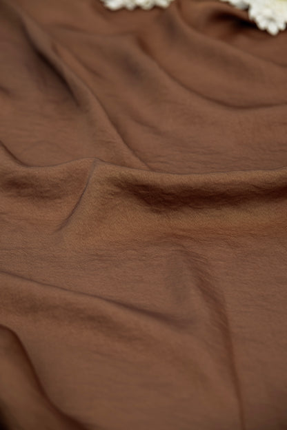 A close-up of draped brown fabric with soft folds and a small portion of a white textured trim visible at the top edge.