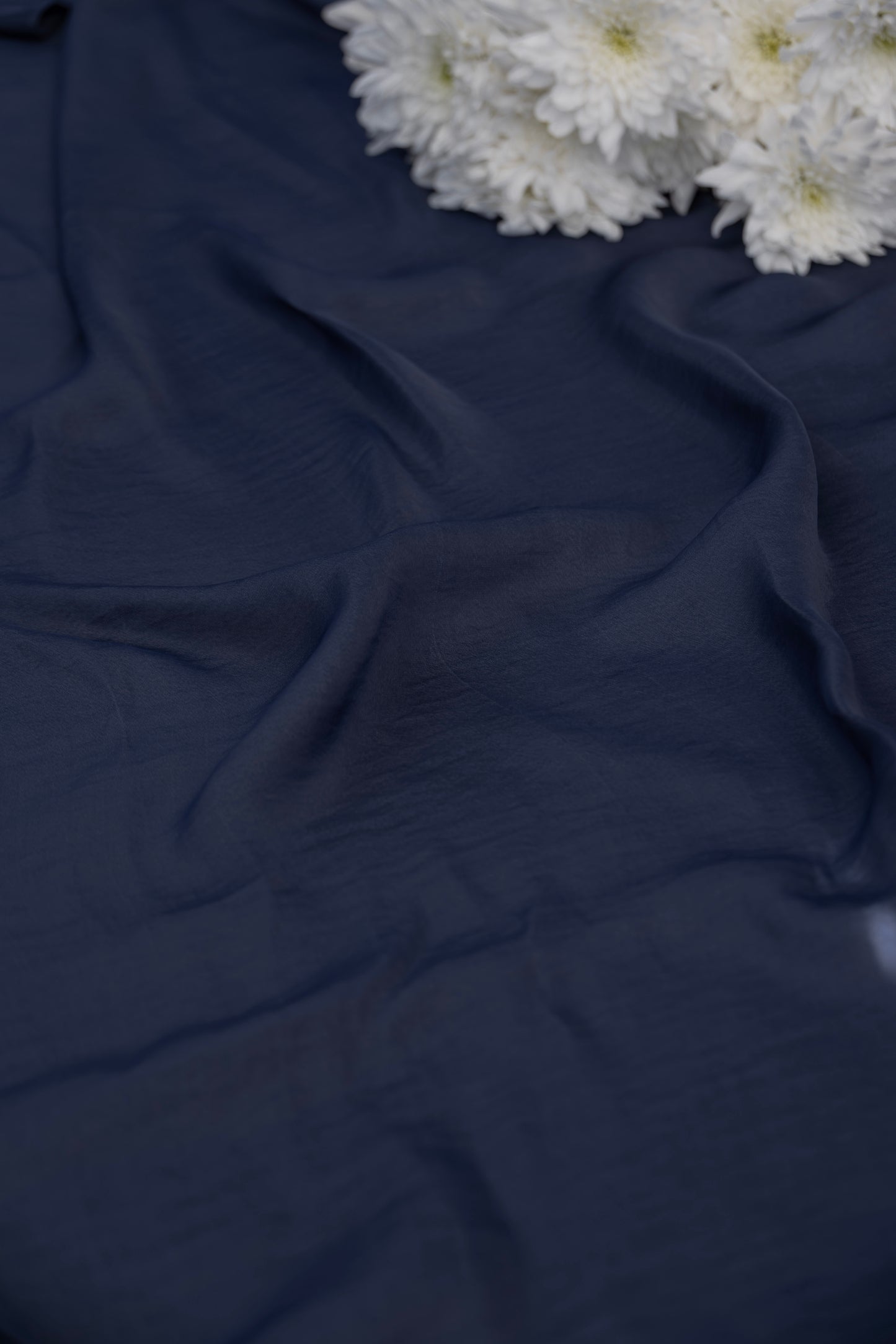 A close-up of draped dark navy blue fabric with soft folds, styled with a small cluster of white flowers in the top right corner.