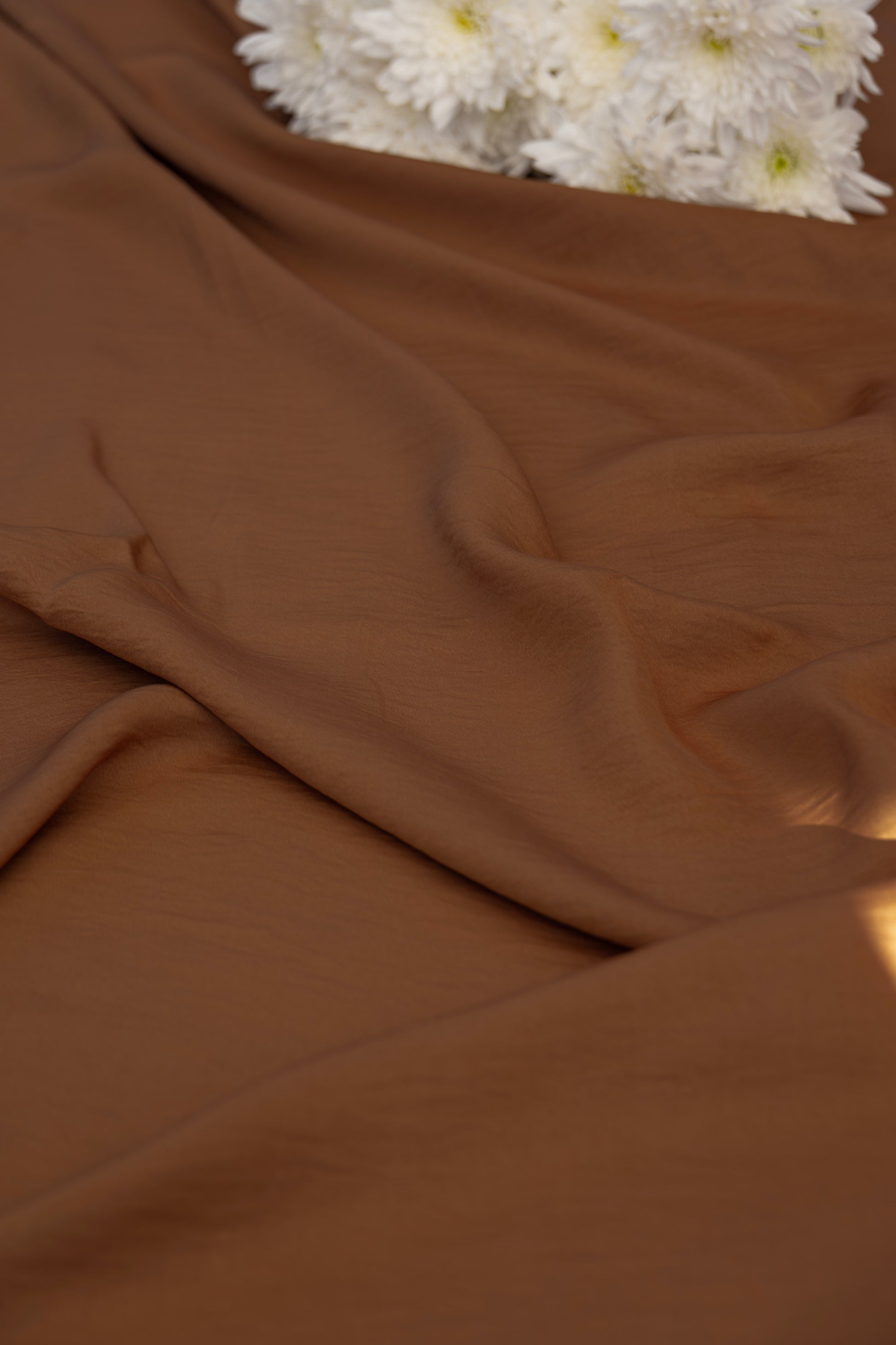 A draped piece of smooth, solid brown fabric with soft folds, styled with a small cluster of white daisies in the top right corner.